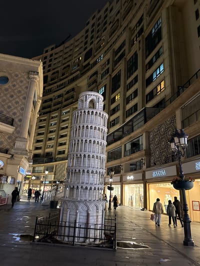 This nighttime image captures a pedestrian area in Esenler, Turkey. A detailed, light-colored replica of the Leaning Tower of Pisa stands prominently near the center-left, encircled by a low, dark metal fence. The tower features multiple arched tiers and exhibits a noticeable lean.

The scene is flanked by multi-story buildings under a dark sky. To the left and curving into the background, a tall, light-toned building with numerous windows, some illuminated, dominates. To the right, another building houses ground-level retail establishments with large glass display windows, revealing merchandise. Visible store signage includes "KAYRA" and partial views of "Flor" (likely Flormar) and "O GIE". Above these storefronts, decorative geometric patterns adorn the building facade.

The ground is paved and shows several reflective puddles, suggesting recent rain. Multiple ornate, dark-colored street lamps with multi-pronged lights illuminate the pathway; some include hanging planters. Approximately 10-12 individuals are present on the pedestrian path, walking in various directions, singly or in pairs. Their attire is casual, and their faces are generally indistinct due to distance and ambient lighting. No specific interactions are discernable among the pedestrians; they appear to be traversing the shopping or public area.