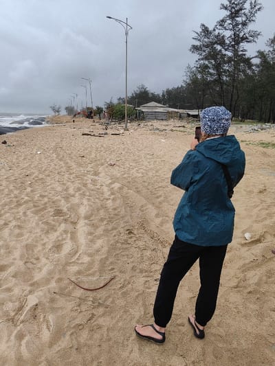 The image captures a sandy beach in Thuận An Ward, Vietnam, under an overcast, grey sky. One individual is visible from behind, standing on the light-brown sand in the mid-ground. This person wears a teal-blue waterproof jacket, black pants, and black open-toed sandals. A blue and white patterned beanie covers their head, with light-colored hair visible beneath. A dark strap from a bag crosses their right shoulder. Both hands firmly hold a dark-colored smartphone horizontally, aimed forward, suggesting they are photographing or videoing the scene.

The immediate sandy ground exhibits numerous footprints and scattered small debris. A distinct reddish-brown bent stick or natural object, resembling a miniature bow, lies on the sand in the lower-left foreground.

In the background, the ocean is visible to the left, with grey waves breaking against dark rocky formations and the shoreline. A series of at least six streetlights, each with a double-arm fixture, extends along a path or road parallel to the beach. Further down the beach, an orange excavator is partially visible. Behind the line of streetlights and trees, several simple, single-story structures with corrugated metal roofs and light-colored walls are situated amongst denser green trees.