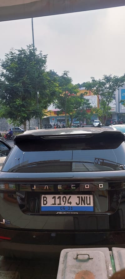 The image shows the rear of a black SUV parked on a street in Cijantra, Indonesia. The car has a license plate that reads "B 1194 JNU" and "06-31" underneath. The SUV has the letters "J A E C O O" embossed on its rear. In the background, there are trees, buildings, and parked motorcycles. The sky is overcast. The focus is on the rear of the vehicle and its license plate.