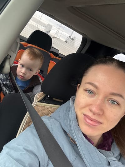 A selfie taken inside a car. A woman with light skin, green eyes, and brown hair is wearing a light blue jacket and a seatbelt. Behind her, a young boy with light hair is sitting in a car seat, also secured with a seatbelt. The background shows a building with multiple windows. The location is Lipaki, Russia.