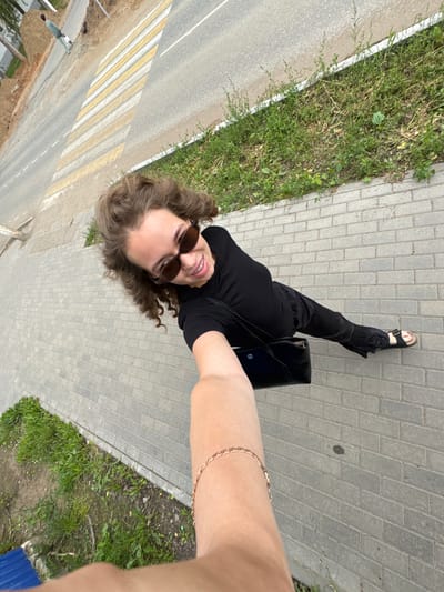 A woman with curly brown hair and sunglasses, wearing a black top and black pants, takes a selfie while standing on a paved sidewalk. She is holding a black purse. In the background, there is a road with a pedestrian crossing. The scene appears to be outdoors in an urban or suburban setting, likely in Chaikovsky, Russia, given the context.