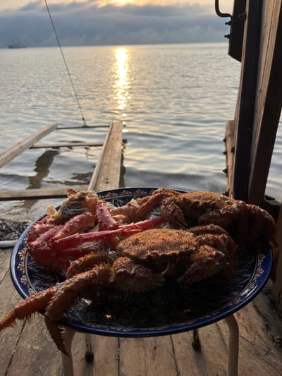 The image shows a plate of cooked crabs on a wooden surface by the water. The crabs appear to be a mix of red king crab legs and hairy crabs. In the background, the sun is setting or rising over the water, casting a golden reflection. There is a wooden ramp or ladder leading into the water, and a ship is visible in the distance. The scene is set at dusk or dawn. The location is identified as Sovetskaya Gavan', Russia, which is a port city on the Tatar Strait.