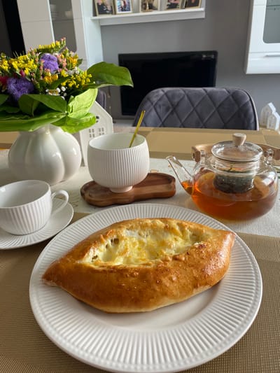The image depicts a breakfast setting on a table, possibly in a home. In the foreground, a white, fluted plate holds a golden-brown pastry filled with melted cheese. To the left, a matching teacup and saucer are positioned. Behind these, a white, ribbed ceramic cup sits on a wooden coaster, and next to it, a glass teapot filled with amber-colored tea and a tea infuser is placed. A bouquet of colorful flowers in a white vase adds a decorative element to the left side of the scene. In the background, a blurred image shows a gray quilted chair, a television screen, and shelves with framed pictures, suggesting an indoor environment, possibly a dining room or living area. The table is covered with a beige placemat. There are no people visible in the image. The location of Alzey, Germany is not evident from the image content.