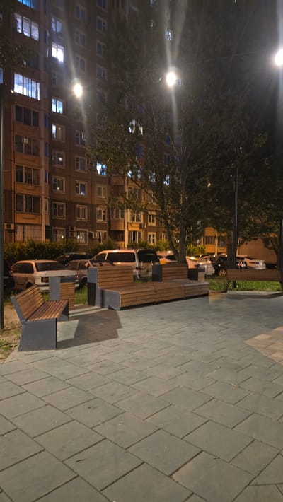 The image depicts a nighttime scene in a residential area, likely a courtyard or public space, in Razvilka, Russia. In the background, a tall, multi-story apartment building is visible with numerous windows illuminated by interior lights, suggesting that it is inhabited. Several trees with dark foliage are positioned in front of the building and extend into the foreground, partially obscuring the structure.
In the middle ground, a row of parked cars is visible. These appear to be a mix of sedans and vans, with headlights and taillights indicating their presence. Some of the cars are partially obscured by trees and the lighting conditions.
In the foreground and mid-ground, there is a modern seating area with wooden benches and dark-colored metal supports. The benches are arranged in a modular fashion, with some sections featuring a slatted wooden design and others appearing solid with perforated metal detailing. A paved walkway, constructed with large, rectangular gray tiles laid in a staggered pattern, occupies the immediate foreground.
Streetlights are present, casting a bright glow that creates lens flare effects in the image. The overall lighting is dim and atmospheric, characteristic of nighttime urban environments. There are no visible people or animals in the scene, and no discernible actions or interactions are taking place. The scene appears to be a quiet, well-maintained outdoor public space.
