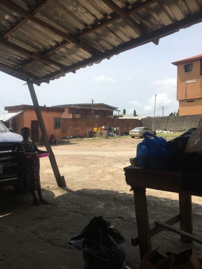 The image captures an outdoor scene in Awjaw, Nigeria, under a corrugated metal roof supported by wooden beams. In the shaded foreground, a young individual in a patterned dress stands barefoot near a white vehicle on the left, holding a pink tray containing white objects. A black plastic bag rests on the ground in the mid-foreground. To the right, a wooden table supports a blue bag and other dark items, with cardboard boxes visible underneath. The sunlit midground consists of an unpaved dirt area. Two individuals are seated near a peach-colored building on the left, where various clothes hang on a line against a reddish-brown wall. A light-colored sedan is parked in the right-midground, near a low concrete block wall and a utility pole. The background features a multi-story reddish-brown building on the far right, and another building with a dark roof behind the left-midground structures. The sky is light blue with scattered white clouds.