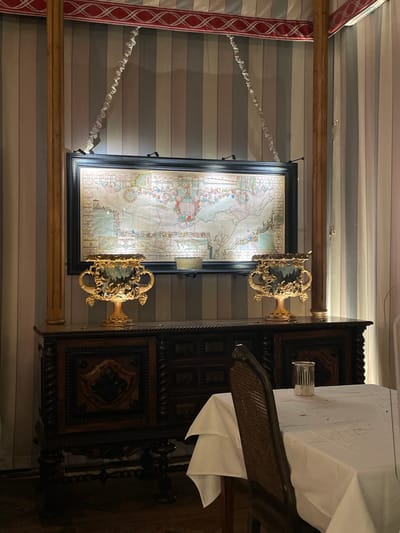 This image depicts an interior scene, specifically a dining area, located in Narbonne, France. The rear wall is covered with vertical stripes in alternating shades of light gray, medium gray, and white, bordered at the top by a decorative red and white patterned fabric. Two fluted wooden pillars flank a central display. A large, horizontally oriented map, featuring an antique style with coastal details and ornate cartouches, is presented in a dark, possibly black, frame and illuminated by two mounted spotlights positioned at its top edge. Below the map, an ornate dark wood credenza, characterized by intricate carvings including twisted columns and diamond patterns, supports two symmetrically placed, golden, internally illuminated urns adorned with grape clusters and foliage. Two decorative light-colored textile or beaded swags extend diagonally from the upper wall border to the top corners of the map frame. In the foreground on the right, a dining table is partially visible, draped with a white tablecloth exhibiting minor dark discolorations. A dark brown chair with a woven cane back is positioned at the table, next to a small, white, cylindrical holder with vertical slots, likely for napkins or condiments. A translucent partition is partially visible on the extreme right edge of the frame. No individuals are present, and no actions are depicted within the static scene.