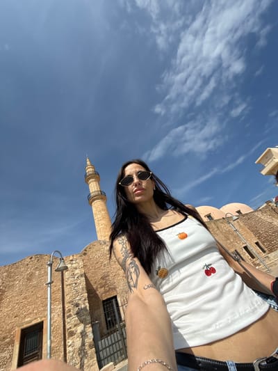 A low-angle, medium shot captures a woman taking a selfie in Rethymno, Greece. The subject, adorned with sunglasses, is positioned in the foreground, framed by a blue sky with light clouds, and the minaret of the Neratze Mosque. The building is constructed of stone. She is wearing a white shirt with embroidered fruit. The building behind the woman has small windows and arched details. A streetlamp is attached to one of the buildings, and a metal grate can be seen covering a window. The composition is oriented on its side.
