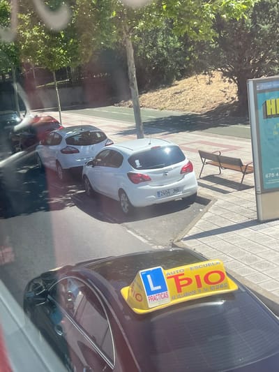 The image captures a street scene in Salamanca, Spain, on a sunny day. In the foreground, a dark sedan with a yellow driving school sign on its roof is parked. The sign displays "AUTO - ESCUELA" and "PIO" in large red letters, along with "PRACTICAS" and a phone number. Behind the sedan, several other cars are parked, including a white hatchback with the license plate "2404 JNB," and another white car. The cars are parked along a sidewalk lined with trees casting shadows on the pavement. In the background, a billboard with partially visible text and numbers can be seen. The overall impression is that of a typical urban street with parked vehicles and pedestrian infrastructure.