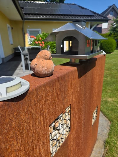 The image shows a rustic, weathered Corten steel wall in a garden setting. A small, terracotta bird figurine sits on top of the wall, next to a modern-looking, silver bird feeder shaped like a house. The wall has two square openings filled with white stones. In the background, a yellow house with solar panels on its roof is visible, along with a green lawn and some trees. The scene appears to be a residential garden. The image was taken in Dissen am Teutoburger Wald, Germany.
