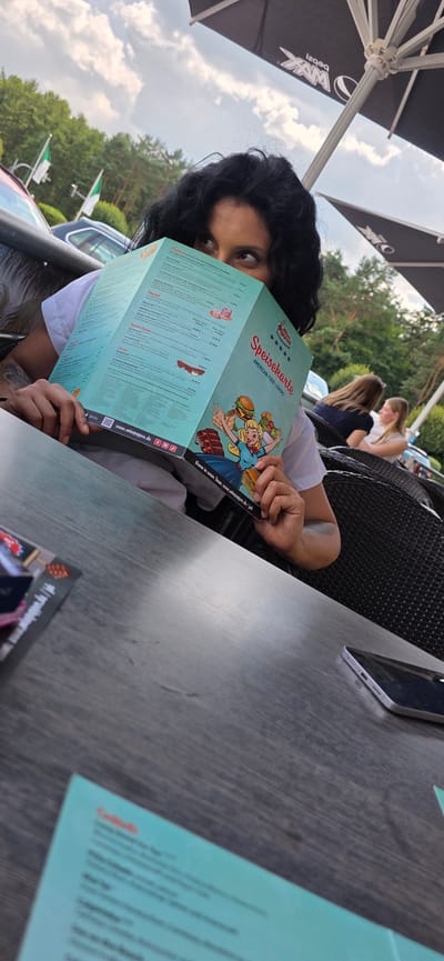 A woman with dark, curly hair is holding a turquoise menu that reads "Speisekarte American Food Lovers" in front of her face. She is wearing a white t-shirt and is seated at a dark wooden table, partially obscuring her face with the menu. The background shows an outdoor seating area with wicker chairs, trees, and a cloudy sky. Two other women are visible in the background, seated at a table. A smartphone is on the table in the foreground. The scene appears to be at "Miss Pepper" in Wietzendorf, Germany, as indicated by the menu and a visible website address on the menu.