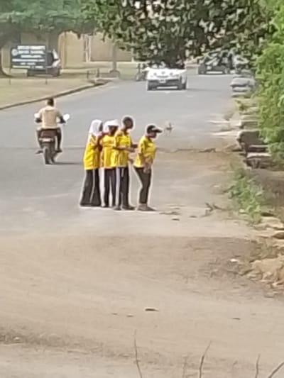 The image captures an outdoor scene on a bright, sunny day, featuring a road bordered by trees and foliage. In the midground, a group of four individuals is standing on the shoulder of the road, which appears to be unpaved or dirt, adjacent to the main paved section. All four are wearing bright yellow short-sleeved shirts.

From left to right within the group:
1.  The first person wears a white head covering, possibly a hijab, and dark, loose-fitting trousers or a long skirt.
2.  The second person is also wearing a yellow shirt and dark trousers, with a white head covering, possibly a cap or hood.
3.  The third person wears a yellow shirt and dark trousers.
4.  The fourth person, positioned slightly to the right, wears a dark baseball cap, a yellow shirt, and dark trousers. They are slightly turned, seemingly looking towards the right or down.

Behind this group, on the paved section of the road, a person is riding a motorcycle away from the viewer, heading towards the left side of the frame. The motorcyclist is wearing a light-colored, possibly beige or white, shirt.

In the background, the paved road continues, and several vehicles are visible in the distance. A light-colored car (possibly white) is visible further down the road, and other less distinct vehicles are also present. On the far left side of the background, partially obscured by trees, a dark rectangular sign with some white text is visible. However, the text is too blurry and distant to be legible. The road appears to have a well-defined paved section in the center, with unpaved or less maintained shoulders on either side. Green trees and dense foliage line both sides of the road, suggesting a semi-rural or park-like setting. The overall image has a slightly soft focus.