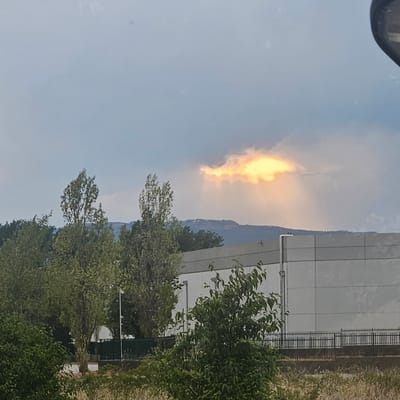 The image shows a bright patch of light illuminating a cloud in an overcast sky, creating a dramatic effect against the muted tones of the surrounding sky and landscape. Below the sky, a line of trees runs horizontally across the frame, with a large, modern building situated behind them. The building has a light-colored facade and a dark, reflective roofline. A black metal fence runs along the front of the building, separating it from a grassy area in the foreground. The overall scene suggests an industrial or commercial area on the outskirts of a city. The location is the city of Vic, Spain.