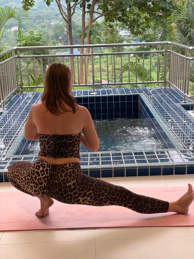 A woman wearing a leopard print tube top and leggings is performing a yoga pose on a pink mat on a tiled patio. She is facing away from the camera, with her legs spread wide and her arms in a prayer position at her chest. Behind her is an elevated, tiled hot tub with bubbling water. The patio overlooks a lush, green hillside with trees and houses in the distance, suggesting a location in Ban Kao Dua, Thailand. A metal railing borders the patio. The sky is overcast, indicating a humid or rainy day.