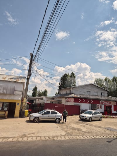 This image captures a street scene in Răcăciuni, Romania, on a sunny day with scattered clouds. In the foreground, a paved road is visible, with a slightly raised curb and a section of concrete with a patterned texture. A light-colored sedan is parked on the right side of the frame, and another similar car is parked further to the left. A man in a green long-sleeved shirt and dark pants is walking towards the left side of the image, near the parked cars.

To the left, a building with a corrugated metal roof and white siding is present. The building has a yellow-painted entrance area with signs and a wooden bench outside. A leaning utility pole, heavily laden with numerous electrical wires and junction boxes, is prominently situated in the middle left of the frame, obscuring part of the sky.

In the background, another building with a light-colored facade and a red awning is visible, bearing the "PROFI LOCO" logo. Several stacks of tires are displayed next to this building. Trees with dense green foliage line the background, suggesting a semi-urban or suburban environment. The overall scene depicts a commercial area with visible infrastructure and everyday activities.
