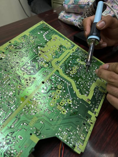 The image displays an individual performing electronics repair on a large, green printed circuit board (PCB) in an indoor setting identified as San Antonio, Panama. A person's right hand, with light brown skin, holds a light blue and black soldering iron, its tip applied to a solder point on the PCB. A partially visible left hand provides support to the board. The PCB, positioned on a dark brown wooden surface, features numerous silver solder pads and green conductive traces. In the background, a metal coil stand designed for a soldering iron is visible, along with a blurry, patterned fabric object and a dark grey garment worn by an unidentifiable individual.
