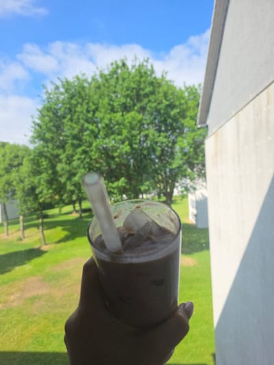A hand holds a glass of iced coffee with a straw in front of a window. Outside the window is a view of a green tree, a blue sky with scattered clouds, and a green lawn with a fence in the distance. The location is Oyten, Germany.