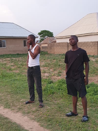 Two young Black men stand outdoors in Bana Stream, Nigeria. One wears a white tank top and black jeans and appears to be talking on the phone, while the other wears a black t-shirt and black shorts. They are both standing on a grassy area next to buildings. One building is covered in corrugated metal, and the other is made of bricks. A dirt path is visible in the grass.
