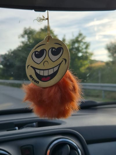 This media file shows a close-up of a car air freshener hanging from a string. The air freshener is circular with a cartoonish smiley face. The face has large, wide eyes, a wide grin showing teeth and a pink tongue, and a small circular nose. The air freshener has a fluffy, orange pom-pom attached to it. The brand name "areon" is visible in small, black text on the bottom right of the smiley face.
The air freshener is hanging in what appears to be a car's interior, with a portion of the car's dashboard and an air vent visible on the right side of the frame. The background, seen through what is likely the car's windshield, shows a bright sky with some clouds and green trees, suggesting it is daytime and the weather is fair. The overall impression is one of a cheerful and possibly scented vehicle interior. The location context of Osikovitsa, Bulgaria, does not provide specific visual cues within the image itself. The image captures a moment of everyday life inside a vehicle.