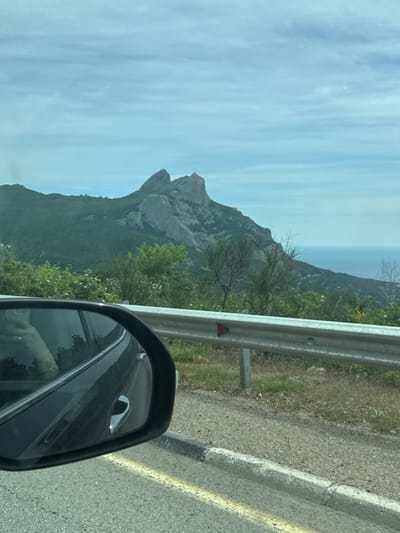 The image depicts a landscape scene, seemingly captured from inside a vehicle, possibly a car, due to the presence of the car's side mirror. The focal point of the image is a mountainous terrain, possibly part of the landscape near the city of Орлиновский округ, Ukraine, as suggested by the location tag. The mountain is mostly covered in green vegetation. Above the mountain is a blue sky with some cloud cover. A roadside safety barrier is also visible on the right side of the image. A portion of the road is visible at the lower right.