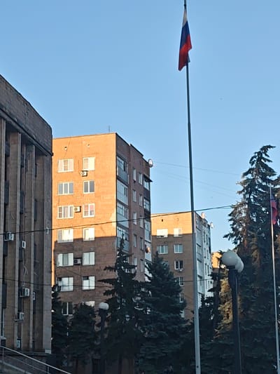 The image depicts a street scene in the city of Chystiakove, Ukraine. A tall flagpole with a Russian flag is prominently featured against a clear blue sky. To the left of the flagpole, a multi-story building with a light brown brick facade and numerous windows is visible. The building appears to be an apartment complex or administrative building. To the left of this building is another imposing structure with a gray, concrete facade and vertical architectural elements. In the foreground, dark green coniferous trees are scattered, partially obscuring the lower portions of the buildings. A lamppost with a distinctive round, multi-bulb light fixture stands near the center-right of the frame. The overall lighting suggests it is daytime, possibly late afternoon or early morning, given the shadows cast. There are no people visible in the image. The environment appears urban with evident architectural structures and vegetation.