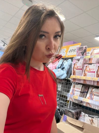 A young woman with long brown hair and full makeup is posing for a selfie in a supermarket aisle in Nurkeevo, Russia. She is wearing a bright red t-shirt with a silver heart-shaped pin on it. The background shows shelves stocked with various products, including what appear to be hosiery and slippers. A price tag with "499" is visible on a yellow sign. The lighting suggests an indoor retail environment.