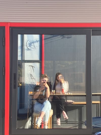 The image shows two people reflected in a window. The window is dark gray with a red frame and is on a building with vertically striped walls. The reflections appear to be of two young women, both holding phones and taking photos. One is wearing light blue pants, white sneakers, and a dark-colored top. The other is wearing a white shirt, and dark pants, and has on pink socks or slippers. The window also reflects signage with script. The location is Satovcha, Bulgaria.