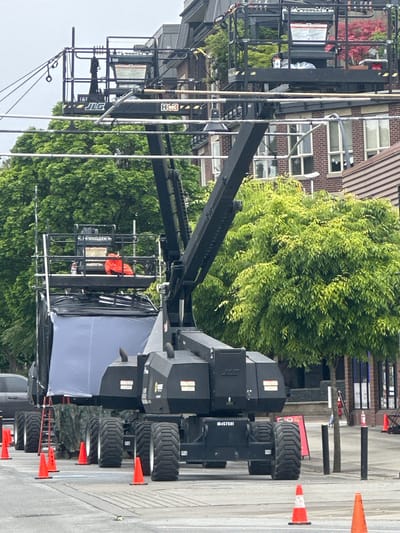 The image depicts a film set in Port Coquitlam, Canada, likely a street scene. A JLG boom lift, a type of aerial work platform, is prominently positioned in the frame. The lift is surrounded by orange traffic cones, indicating a cordoned-off area. A section of a building with multiple windows is visible in the background, partially obscured by a large green tree. Other equipment and people associated with film production are present, though specific details are difficult to discern.