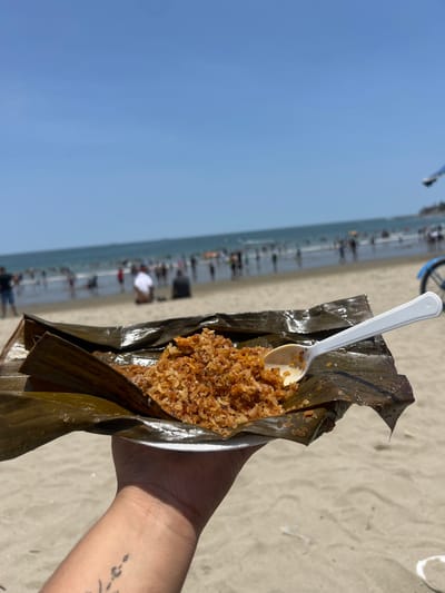 A high-angle forensic shot captures a human's left hand holding a serving of food on a white foam plate, presented in dark green banana leaves, against the backdrop of General Villamil Playas, Ecuador. The food consists of a reddish-brown, granular mixture, likely rice or a similar grain, with a white plastic spoon resting on its surface. The hand exhibits a faded, dark tattoo on the inner forearm. In the blurred background, a wide, sandy beach is visible, populated by numerous individuals. The ocean, light blue in hue, extends behind the beach, with many people wading and swimming in the water. The sky above is clear and light blue. To the far right, a partial blue bicycle or similar object is discernible. The scene is illuminated by bright daylight, casting no discernible shadows.