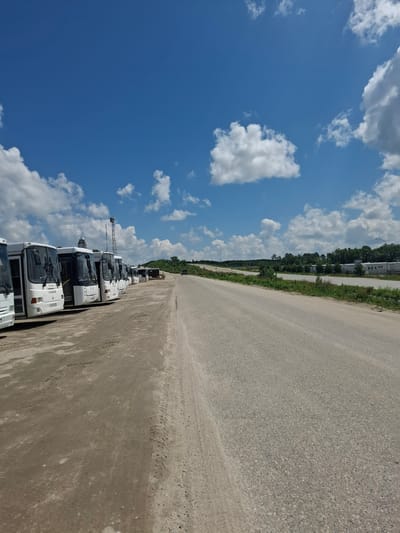 The image depicts an outdoor scene at the Tecnimont camp in Russia. A line of white buses is parked on a dirt lot on the left side of the frame. The buses are oriented parallel to a paved road that stretches into the distance on the right. The road is bordered by sparse vegetation and trees. The sky above is blue with scattered white clouds. A cell tower is visible in the background, behind the buses. There are no people visible in the image. The overall impression is one of a quiet, possibly industrial or transit-related location.