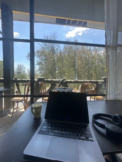 The scene depicts an indoor workspace overlooking an outdoor natural environment in Phra Ae, Thailand. In the foreground, a silver laptop with a dark display is open on a dark, rectangular table. To the left of the laptop is a light yellow mug, and to its right are black JBL headphones. Beyond the table, a large window provides a view of an outdoor terrace area. This terrace features a distinctive railing designed to resemble gnarled tree branches, along with several dark-topped tables and curved wooden chairs. In the background, a dense forest of tall, slender trees extends under a clear blue sky dotted with scattered white clouds, with a narrow body of water visible on the distant horizon. Interior elements include a dark ceiling with exposed structural components above the window, a sheer light-colored curtain on the right, and a thick, dark, textured pillar on the left. No individuals are present in the image.