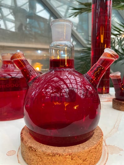 The image depicts laboratory glassware filled with a red liquid. The glassware includes a spherical flask with multiple necks. One neck has a clear glass stopper. Other necks are short and slightly angled. The flasks sit on thick cork stands. The liquid in the flasks is a deep red color and appears to be translucent. The background shows reflections of a building with a grid-like structure, suggesting a window. The lighting is bright and diffused, creating highlights on the glassware. The location is suggested to be Konya, Turkey but there is no supporting evidence of this in the image.