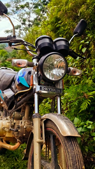 The image displays a close-up, low-angle, front-facing perspective of a utility motorcycle situated amidst dense green vegetation, specified as being in Pung Luong, Vietnam. The motorcycle is a "DETECH" brand, with its name prominently embossed on a chrome badge located at the base of the handlebars, above the front suspension. The vehicle features a black fuel tank adorned with stylized blue and white decals, a black seat, and black handlebars equipped with mirrors and controls. A large, circular headlight with a clear, reflective lens dominates the front, flanked by amber-colored turn signals. Above the headlight are two dark, circular instrument casings. The front fender is a dirty metallic silver-grey, and the lower portions of the front suspension forks, engine block, and front wheel are heavily coated in dried mud and dirt, indicating recent use in rugged terrain. The engine casing appears to be a dull metallic beige, and a segment of a gold-brown exhaust pipe is visible on the left side. The front wheel has a dark rim with a faint red stripe. The background consists of a vibrant, varied assortment of green foliage, including trees and ferns, creating a lush, natural environment under natural daylight. No people or animals are present in the scene.