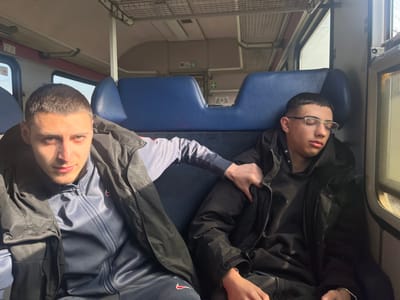 The scene depicts the interior of a train carriage, reportedly located in Ruska Bela, Bulgaria. Two males are seated in blue upholstered seats, illuminated by direct sunlight entering from windows on the right.

The male on the left, a young adult with short, dark hair, wears a dark sleeveless puffer vest over a navy blue tracksuit top featuring a white zipper and a small red Nike swoosh. He is looking directly at the camera. His left arm extends towards the male on his right, with his hand resting on the other individual's upper right arm or shoulder.

The male on the right, a younger individual also with short, dark hair, wears light-framed eyeglasses and a black hooded puffer jacket. He appears to be asleep or resting, with his eyes closed and his head tilted against the seat or window frame. A thin, light-colored bracelet is visible on his left wrist.

The train interior features light-colored wall panels, an overhead metallic luggage rack, and multiple windows. A green emergency exit sign with an arrow and a figure, as well as a "WC" sign, are visible on the far wall of the carriage.