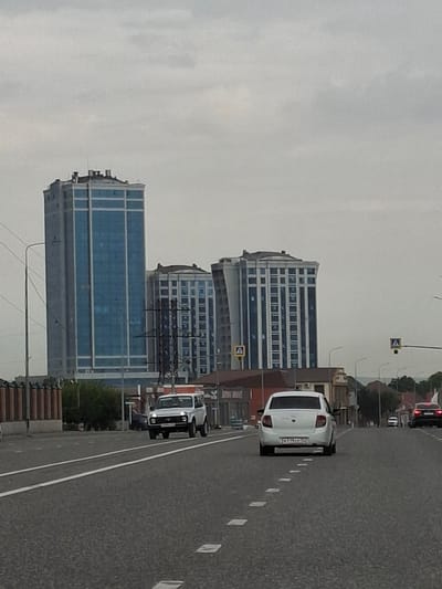 This is a vertical, eye-level, medium shot of a city street in Argun, Russia, on a cloudy day. The scene is dominated by two modern, multi-story buildings with blue glass facades and a gray overcast sky. The buildings are contemporary in design, with repeating patterns of windows. In the foreground, a gray asphalt road with white lane markings stretches into the distance. Several vehicles are visible on the road, including a white car with its license plate visible, and a white van with its headlights on. The overall mood is somewhat subdued due to the cloudy weather. There are no people clearly visible in the image. The time of day appears to be daytime, but the overcast conditions obscure the direct sunlight.