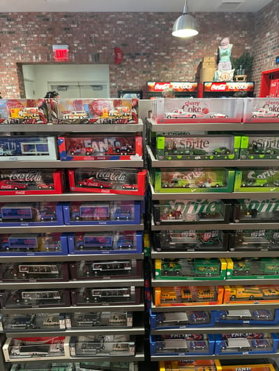 The image shows a display of Coca-Cola and other soda-branded diecast delivery truck toys, arranged on shelves in a retail setting in Lake Buena Vista, Florida, United States. The trucks are packaged in clear plastic and cardboard containers, with the brand names "Coca-Cola," "Cherry Coke," "Sprite," and "Fanta" prominently displayed. The shelves are gray metal. In the background, there is a brick wall, a cooler with illuminated Coca-Cola advertisements, and other merchandise. A doorway is visible with light streaming in from the outside. The lighting is a mix of overhead and reflected light from the merchandise.