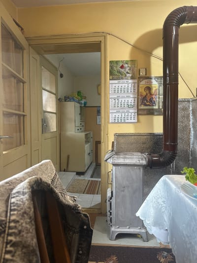 The image depicts an indoor scene within a residential building in Letnitsa, Bulgaria, as indicated by a wall calendar displaying "ОБЩИНА ЛЕТНИЦА" and dates for 2026. The environment features warm yellow-painted walls.
In the foreground, to the left, a gray acid-washed denim jacket is draped over a chair, with a partial view of its texture and buttons.
The midground is dominated by a gray solid fuel stove, positioned on a dark patterned rug. A dark brown, corrugated stovepipe extends vertically from the top of the stove, curving sharply to the right and disappearing out of frame. Behind the stove, the wall is partially covered by a silver, textured heat-resistant panel. To the right of the stove, a table is partially visible, covered with a white tablecloth featuring a floral or damask pattern. On the table, a plate containing green leafy vegetables is visible.
The back wall displays two calendars for the year 2026. The left calendar shows "МАЙ 2026 МАУ" (May 2026) and a picture of a white horse with a red blanket, along with visible dates for March, April, and May. The right calendar features an icon of an angel and "2026" in Cyrillic. Between the calendars, a small, light-colored square object, possibly a light switch or thermostat, is affixed to the wall. A white electrical cable runs horizontally along the upper portion of the wall.
To the left, a light-colored wooden doorway with multiple frosted glass panels leads into an adjoining room. A metallic door handle is visible on the right door of this frame. The adjacent room has light-colored tiled flooring and features two stacked, light-colored modular cabinets against a wall. Various items, including a green pitcher and cardboard boxes, are visible on top of the upper cabinet. A second doorway is visible in the deeper background of this adjacent room.
No individuals are present in the scene.