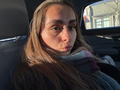 A woman sits in a car wearing a scarf and looking at the camera with slightly pursed lips. Light streams in, creating shadows and highlights on her face and hair. Outside the car window, buildings are visible, including one with an "OPEN" sign in the window. The car's headrest is visible behind her.