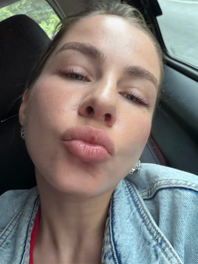 This is a close-up, selfie-style portrait of a young woman. She is angled towards the camera, with her face filling most of the frame. Her eyes are partially closed, and her lips are slightly pursed, giving her a relaxed or playful expression. She appears to be wearing makeup, with mascara on her eyelashes and a subtle blush on her cheeks. She has on a light-wash denim jacket with silver chain detailing on the collar, and a glimpse of a red garment beneath. Her hair is light brown and pulled back. The background is blurred, suggesting an indoor setting, possibly a car, with a hint of green foliage visible through a window, indicating it might be daytime. The image was taken in Dračevice, Bosnia and Herzegovina. There is no visible text in the image.