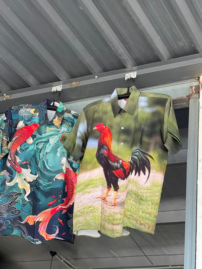 Two shirts are hanging from black hangers against a corrugated metal wall. The top shirt is green and features a photorealistic image of a rooster in a field. The rooster is depicted in vibrant red and black colors, with its tail feathers spread. The bottom shirt is dark blue and features a pattern of koi fish swimming in swirling green water, with stylized clouds and waves. The shirts appear to be new and are displayed outdoors, possibly in a market or shop stall. The lighting suggests it is daytime, and the setting appears to be in an outdoor or semi-outdoor area, given the corrugated metal structure and the visible rust on the frame above. There are no people visible in the image, and the focus is solely on the two garments. The image is taken from a slightly elevated angle, looking down at the shirts.