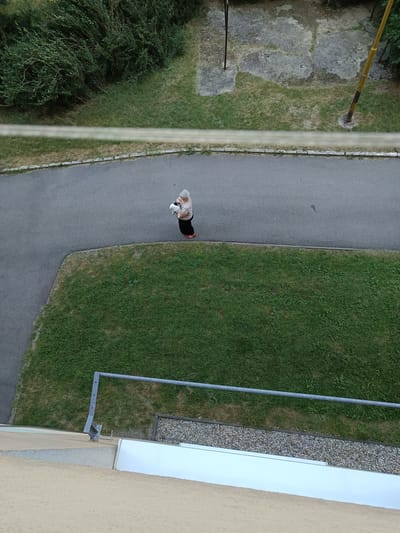 An overhead shot shows an elderly woman holding a small white dog while standing on a paved path. The woman is wearing a light brown shirt and a dark skirt with red shoes. She is in a park-like setting with a patch of green grass to her right and a sidewalk to her left. A metal railing is visible in the foreground, and in the background, there are trees and some concrete structures. This image was taken in Košice, Slovakia.