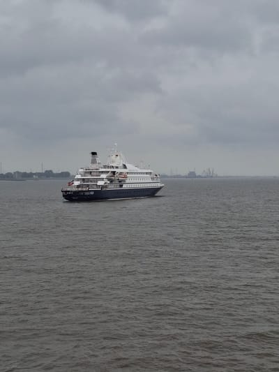 The image shows a cruise ship sailing in water, with land and buildings visible in the background. The sky is overcast. The cruise ship is a dark color with multiple levels and a smokestack. The location is Brunsbüttel, Germany, as evidenced by the general appearance of the coastline and the presence of the industrial structures in the background.