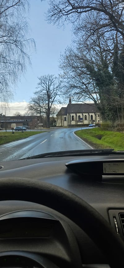 The image, taken from inside a vehicle in Rietberg, Germany, depicts a road leading towards a building resembling a church or chapel. The road appears wet, suggesting recent rain. Trees with bare branches line the left side of the road, contrasting with a patch of green grass on the right. Several parked cars are visible near some buildings along the road. The dashboard and steering wheel of the vehicle are in the foreground, slightly obstructing the full view of the scene. The car's speedometer is visible, displaying values. A small EU flag is seen on a sign in front of the building.
