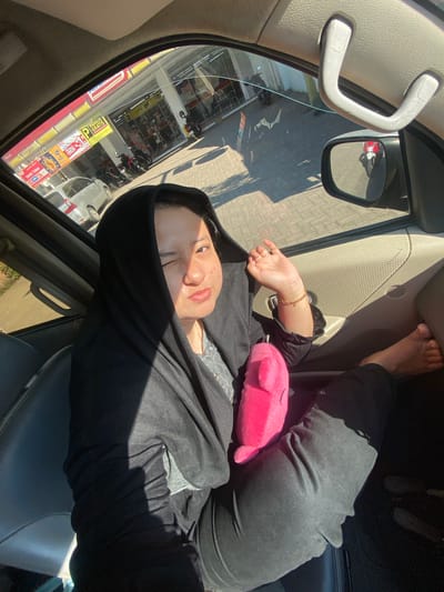 A woman wearing a black hijab and a gray shirt sits in the passenger seat of a vehicle. She is holding a pink plush toy in her lap and has her left arm raised, hand resting on the car door. Her right leg is extended, with her bare foot visible. Sunlight streams into the car, creating shadows on her and the interior. Through the open window, a convenience store with a red and yellow awning and various signs, including one that reads "PARKING GRATIS" (Free Parking), is visible. Several motorcycles are parked outside the store. The ground outside is paved with interlocking blocks. The scene appears to be in Jabon, Indonesia, based on the signage.