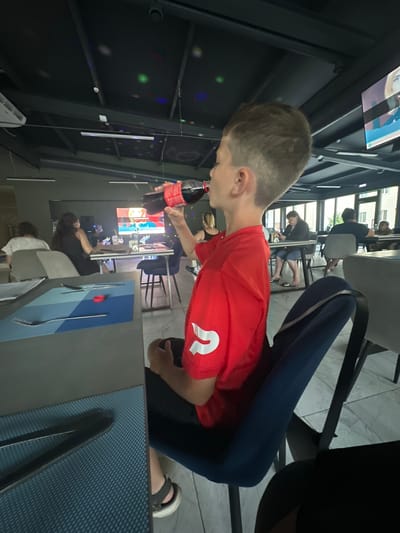In this vertically oriented image taken in Danilovka (Altai Krai), Russia, a young person wearing a red t-shirt with a white logo is drinking from a bottle of Coca-Cola. The person is seated at a table in what appears to be a modern, indoor dining area. They are looking up and to the side, with their arm bent as they bring the bottle to their mouth. The red t-shirt has a prominent white "2" on the chest. The person is wearing dark blue leggings and sandals.
The setting features a dark-colored table with a textured blue placemat and silverware. In the background, other patrons are visible, some seated and some standing. One person in the background is wearing a white shirt and another is wearing a black dress. There is a screen in the background displaying what looks like a cartoon or animation. The ceiling has a black, textured appearance with several small, colored lights that resemble a disco ball effect. The overall ambiance suggests a casual gathering or meal. The lighting is somewhat dim, with artificial lights contributing to the atmosphere. The image seems to have been taken during the daytime, but the interior lighting is prominent.