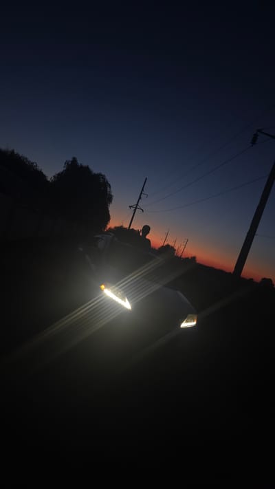 A car's headlights are brightly illuminated, casting strong beams of light across the foreground of the image. The car is positioned at an angle, with its front end partially visible. A silhouette of a person is visible in the driver's seat. The scene is set against a dark, twilight sky with a gradient of deep blue fading to orange near the horizon, suggesting it is either dawn or dusk. Several utility poles with wires are silhouetted against the sky, indicating an outdoor, possibly rural or suburban setting. The surrounding environment is dark and indistinct, with trees and foliage visible as dark shapes against the lighter sky. No specific landmarks identifying Matroska, Ukraine are present in the image.