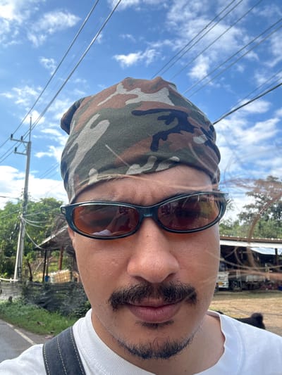 A close-up, chest-level outdoor portrait depicts one individual directly facing the viewer in Ban Mae Na Chalong, Thailand. The person wears a camouflage-patterned bandana, dark-framed sunglasses with amber-tinted lenses reflecting a mobile device, a white t-shirt, a mustache, and a chin strap beard. A dark strap, possibly from a bag or garment, is visible over the left shoulder. The background features a bright blue sky with scattered white clouds and multiple overhead electrical or communication wires connected to a utility pole on the left. Green vegetation is present throughout the middle and background. On the left, behind the utility pole, a rudimentary structure with a corrugated metal roof and stacked objects is visible. On the right, a second structure with a corrugated roof and a white truck are partially obscured. A dark animal is partially visible in the lower right foreground. The scene is illuminated by daylight.