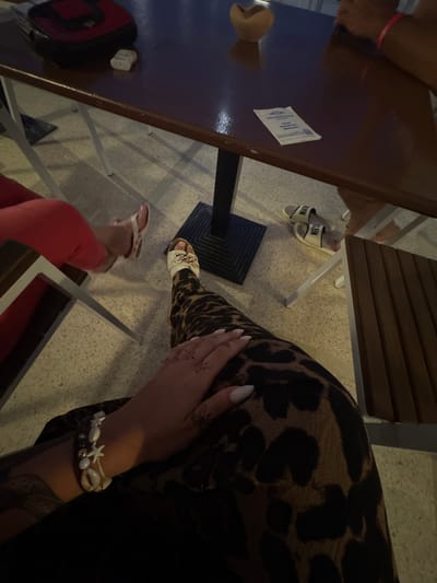 A low-angle, slightly distorted shot depicts a table setting with two individuals present, potentially at an establishment in Mrezga, Tunisia. One person wears animal print leggings and a bracelet made of seashells, and has manicured nails and tattoos. The other individual is wearing pink pants and flip-flops. A pair of sandals with the inscription "KLEIN" is also visible on the floor near the table, which has a brochure or menu on it.