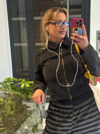 A young adult female is depicted taking a mirror selfie indoors in Nogent-sur-Marne, France. She has blonde hair, wears black rectangular glasses, and white wired earbuds are visible in both ears. She is dressed in a dark, cropped, zippered jacket with a brass-colored zipper and black and grey horizontally striped pants. Her right hand holds a hot pink iPhone, which contains a yellow card visible behind the phone's transparent case, and a silver ring is present on her left ring finger. Her left hand rests on a large green potted plant, wrapped in clear plastic sheeting with visible condensation, situated to her left. In the background, behind her, a dark-framed glass door or window reflects the reversed text "RDC." To the far right, a grey wall panel features a white intercom or control panel. A white shopping bag with yellow ribbon handles hangs from her left side, partially visible. The immediate environment includes a white wall to the left and a tiled or patterned floor.
