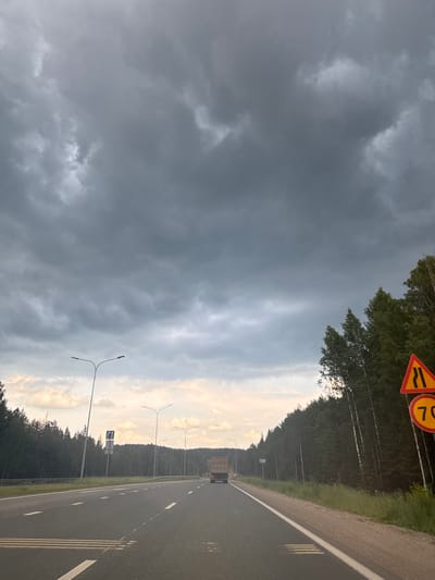 The image depicts a view from a moving vehicle on a paved road, likely a highway, with a dense forest lining the right side and a lighter, sparser forest on the left. The sky is filled with dark, ominous storm clouds. On the right side of the road, there are two traffic signs: one triangular warning sign with a black wavy line on a yellow background, indicating a narrowing of the road, and a circular sign below it with the number "70" in black on a red background, signifying a speed limit of 70. Streetlights are visible along the left side of the road. In the distance, a truck is traveling in the same direction as the viewer. The overall scene suggests an impending storm or the aftermath of one, with a moody and slightly dramatic atmosphere. This image is not definitively located in Nytva, Russia.