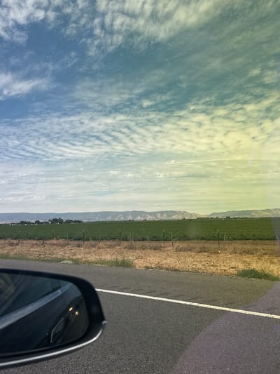 The image depicts a view from inside a vehicle, looking out towards a rural landscape. In the foreground, a portion of a black car's side mirror is visible on the left side of the frame, reflecting the surrounding environment. The road is asphalt, with a white dashed line indicating a lane divider. Beyond the road, a dry, grassy embankment transitions to a vibrant green field. A wire fence with metal posts runs along the edge of the field. In the mid-ground, a range of low, rolling hills can be seen, some with visible vegetation and others appearing more barren. The sky is filled with scattered white clouds against a blue backdrop, with a subtle color gradient from blue to yellow/green, possibly due to lighting or camera effects. The overall scene suggests an open, agricultural area with a wide sky, characteristic of the countryside. The location is identified as Dixon, United States of America.